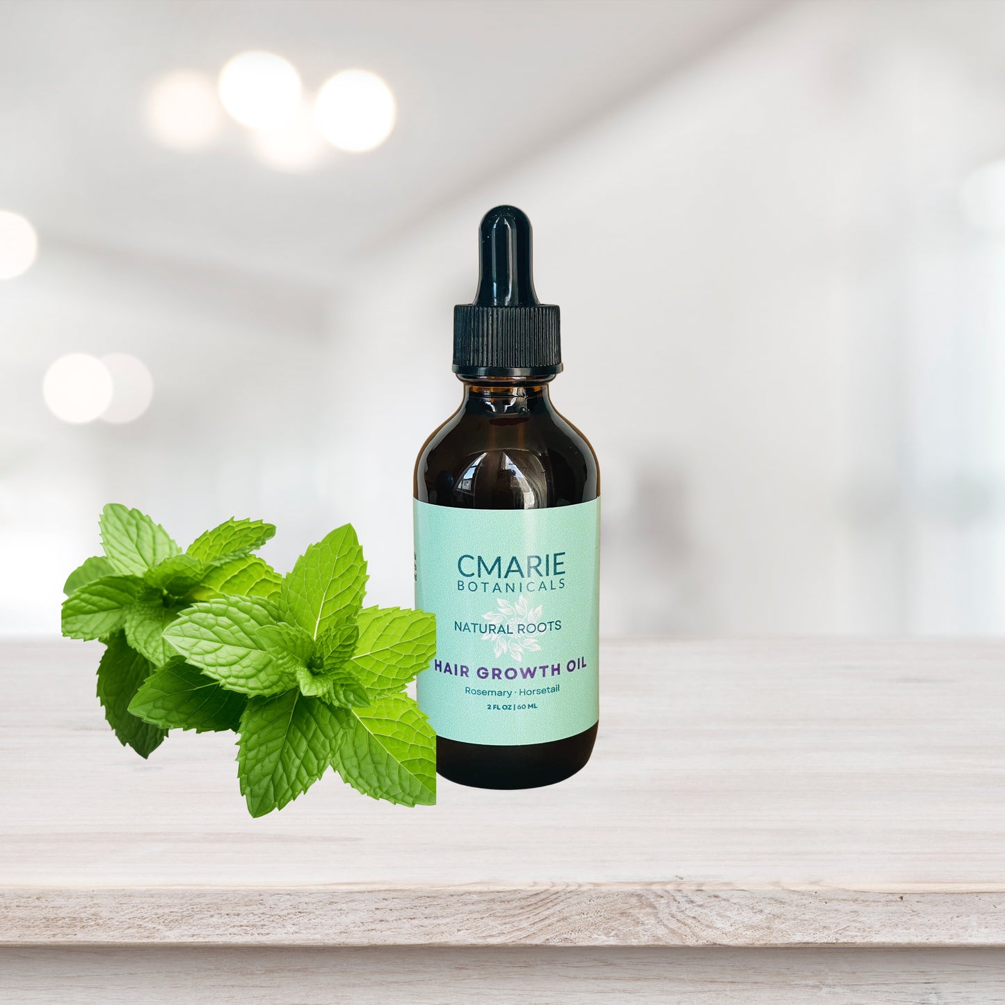 A glass dropper bottle labeled "CMARIE Botanicals Natural Roots Hair Growth Oil" placed on a wooden surface. Fresh peppermint leaves are arranged next to the bottle, with a soft, blurred white background highlighting the product.