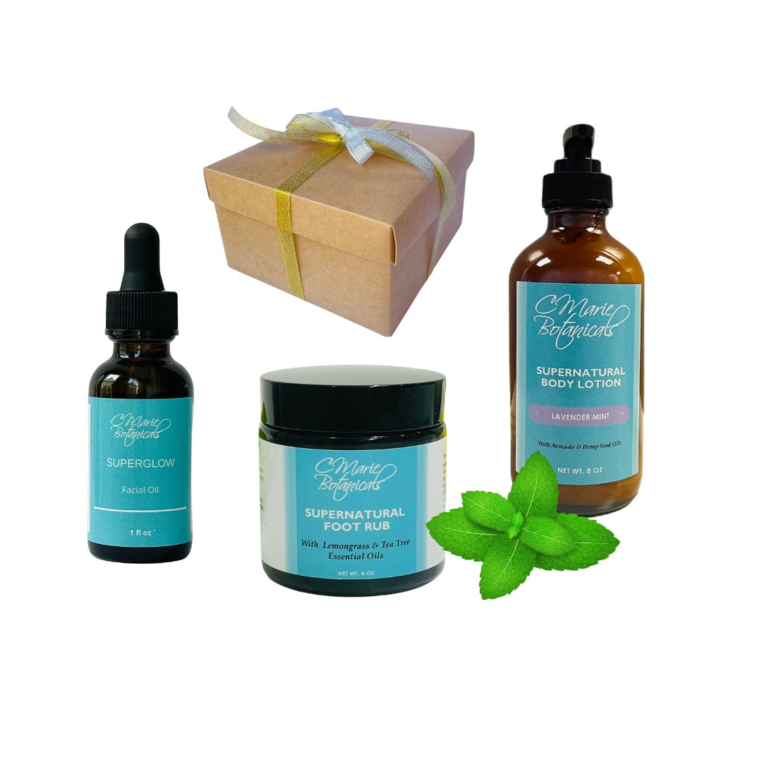 Gift box featuring four natural products: Superglow Facial Oil, Supernatural Foot Rub, with Lemongrass and Tea Tree and Supernatural Body Lotion with a green mint leaf next to it.  The products are arranged next to a brown gift box tied with a gold and silver ribbon.